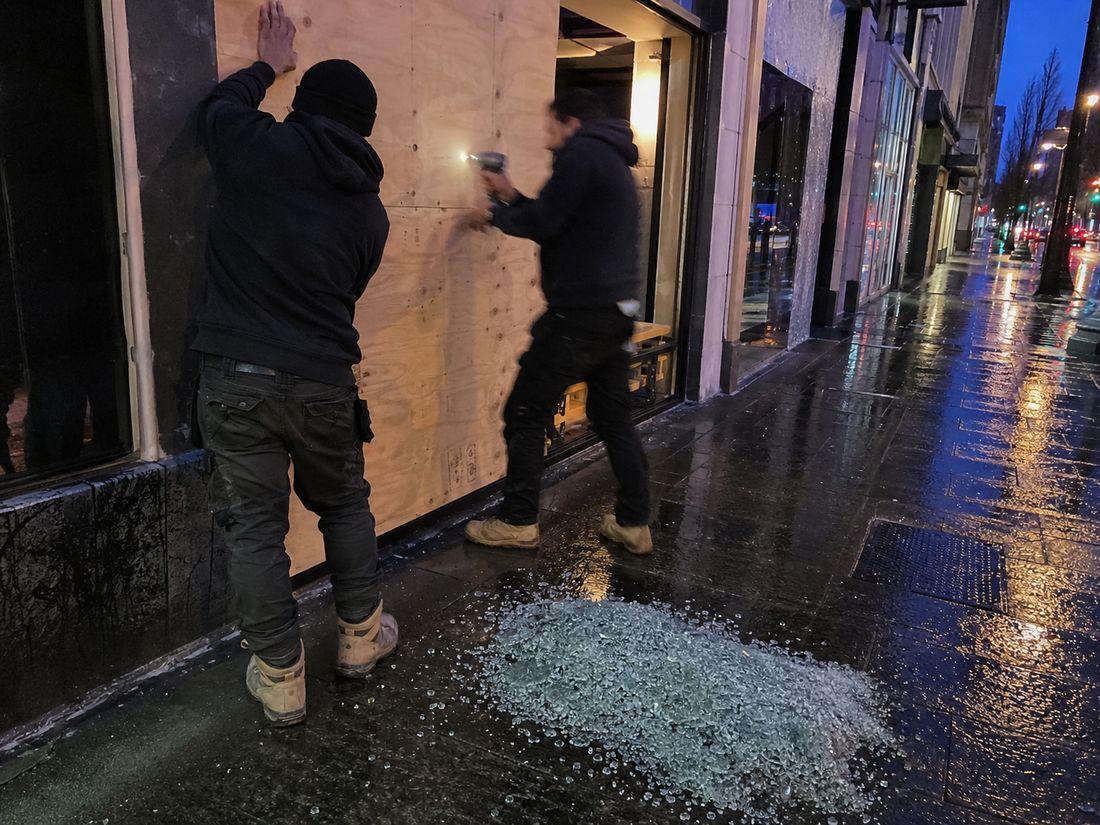Two glaziers screwing a plywood board over a smashed shopfront window at dusk on a wet Wellington street