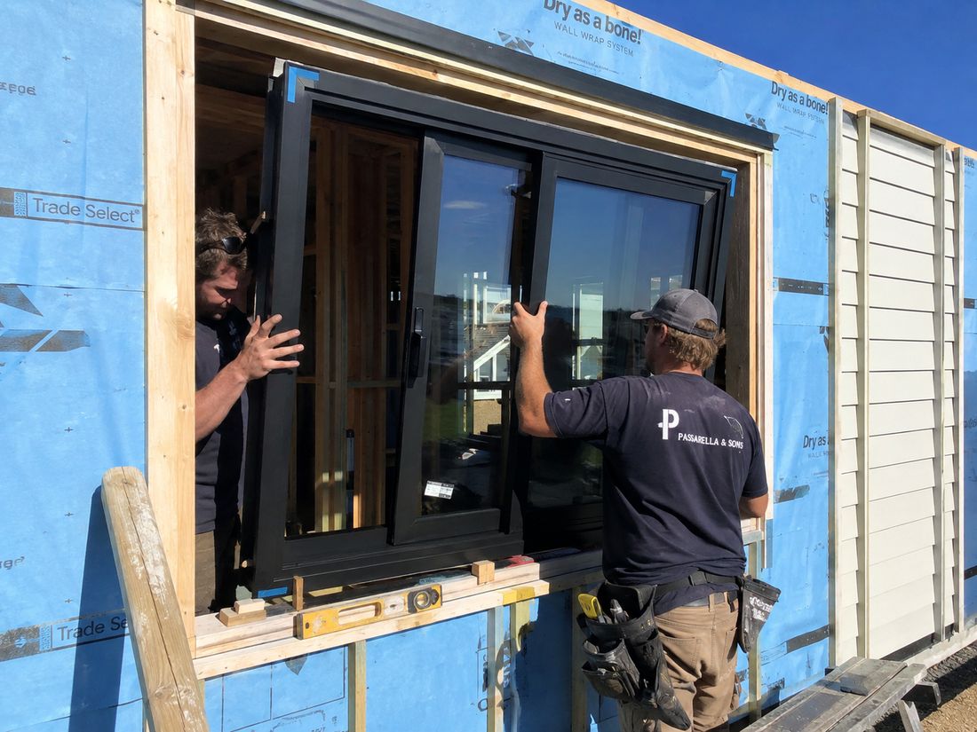 Two installers guiding a new dark aluminium window unit into a framed opening in a wall wrapped in building paper